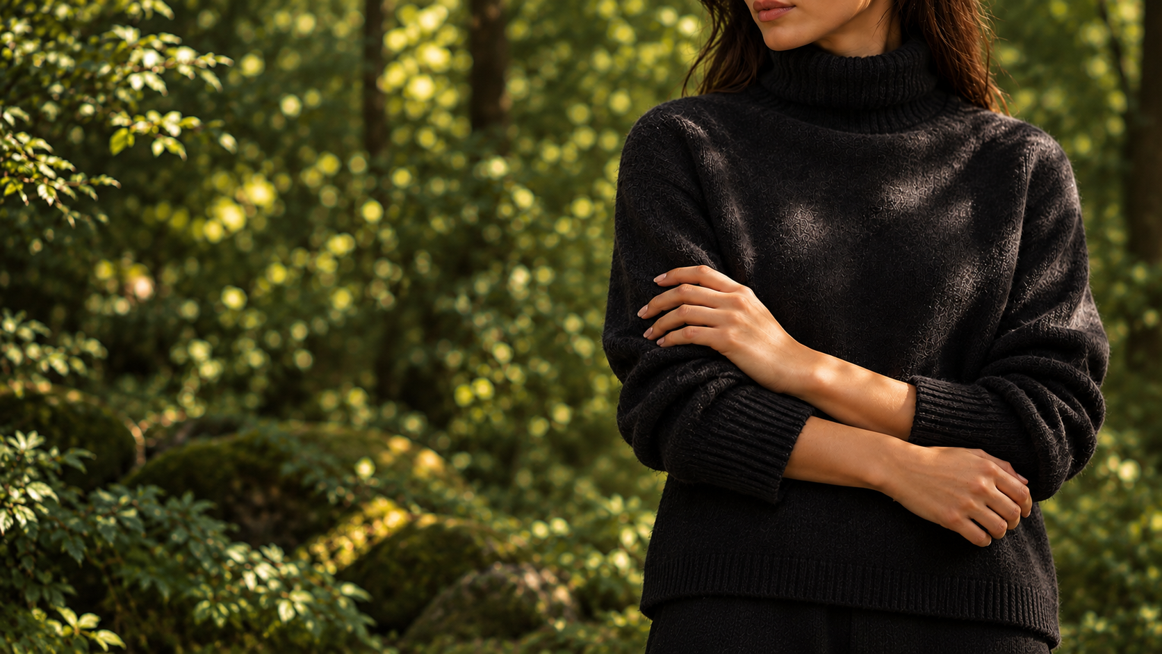 A woman in sculptural black knitwear resting her hands gently on her body in soft woodland light; only lips and chin are visible.